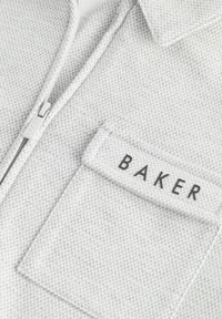 Pelēka iešūta krekla, kas iezīmē teksturētu audumu ar krūšu kabatu, kas apzīmēta ar "BAKER" melnā krāsā. Rāda modernu, ikdienas dizainu.