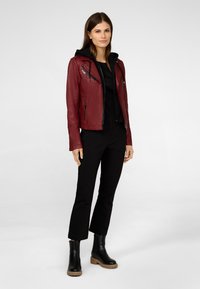 Veste de biker en cuir rouge avec des poches zippées et une couche à capuche noire en dessous, associée à un pantalon noir et des bottes noires épaisses.