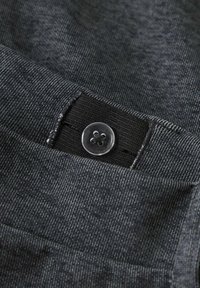 Mørk grå stoff med en fin tekstur, som har en klar knapp og elastisk linning for lukking. Nærbilde av detaljene i plagget.