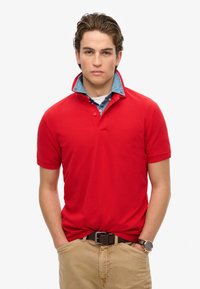 Polo rosso con colletto in denim, maniche corte e patta con bottoni. Indossato con pantaloni beige e una cintura scura. Design semplice e casual.