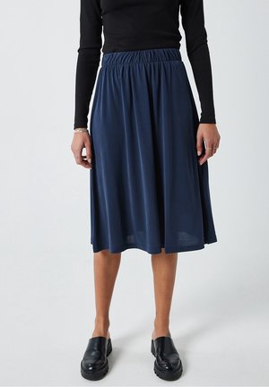 Femme portant une jupe bleu marine longueur genoux avec une taille élastique, un haut noir à manches longues, des chaussures noires sans lacets, et un bracelet et des bagues en argent.