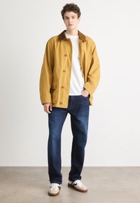 Giacca di tela gialla con colletto in suede marrone, chiusura con bottoni, due tasche frontali, abbinata a jeans blu scuro e sneakers bianche.