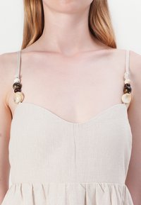 Beige leninki, jossa on muotoiltu vartalo ja röyhelöhame. Säädettävät olkaimet, joissa on koristeelliset helmet eri väreissä.