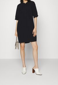 Svart kortärmad klänning med en rynkad halsringning, i kombination med vita öppna tåskor och en liten grå handväska. Enkelt, rent design.