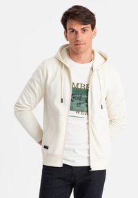 Crèmekleurige zip-up hoodie met een trekkoordcapuchon, voorzakken en zwarte accenten. Draag het over een witte graphic t-shirt en donkere jeans.