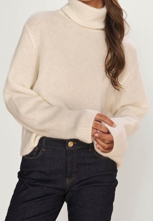 Kermanvärinen neulottu pooloneule, jossa on ribbineulos, rento istuvuus ja leveät hihansuut, yhdistetty tummiin farkkuihin, joissa on kultaiset yksityiskohdat.