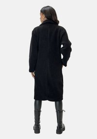 Abrigo largo negro de tela suave y texturizada con un cuello alto, bolsillos laterales y hombros caídos, combinado con leggins negros brillantes y botas.