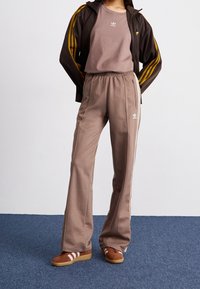 Brown tracksuit with a zip-up jacket and elastic waist pants. Features white stripes and logo accents. Brown shoes with white stripes.