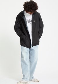 Sort vandtæt jakke med hætte, grå foring, to sidelommer; båret over en hvid grafisk t-shirt og lyseblå jeans.