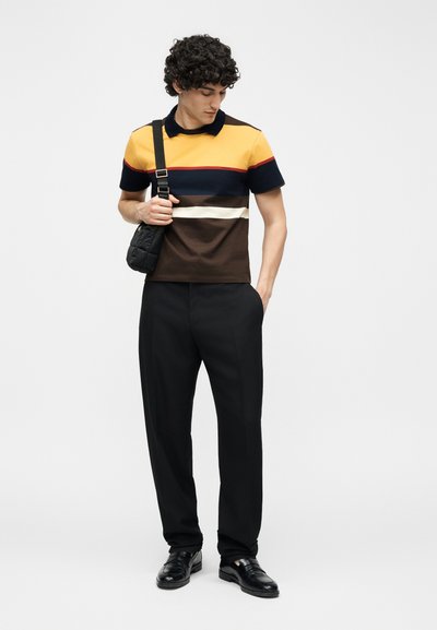 Man wearing a striped yellow, navy, white, and brown polo shirt, black pants, black shoes, and carrying a black shoulder bag.