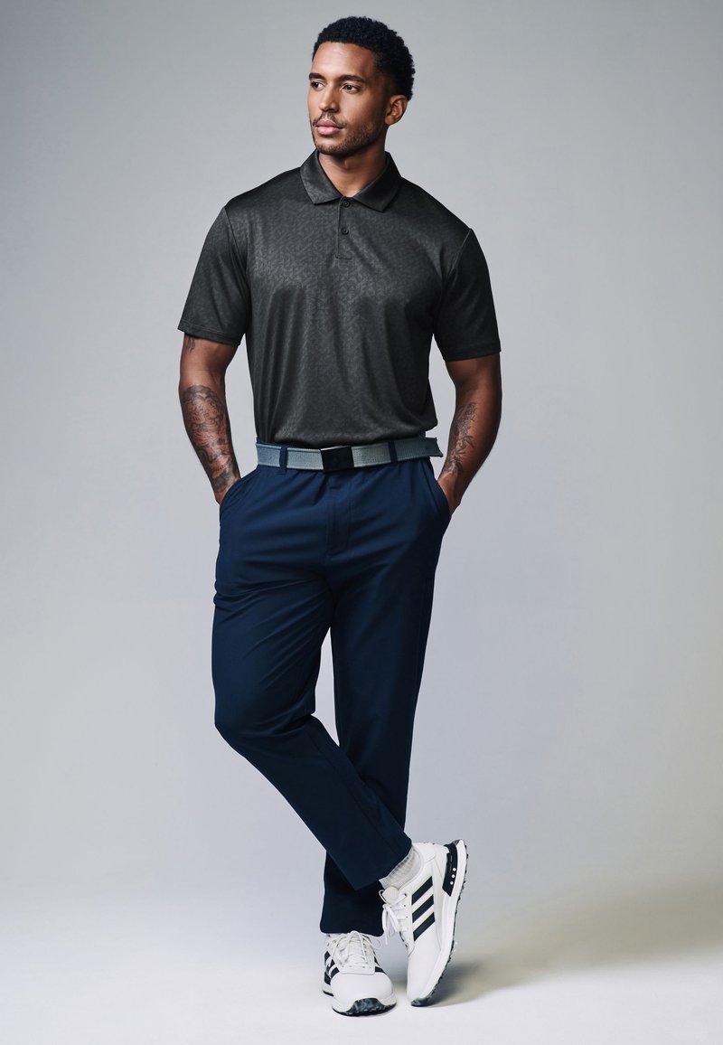 Mann mit Tätowierungen, der ein dunkelgraues Poloshirt, marineblaue Hose, weiße Sneakers und einen grauen Gürtel trägt, steht mit den Händen in den Taschen vor einem grauen Hintergrund.