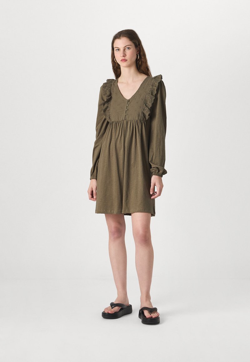 Κακουί φόρεμα με V-λαιμόκοψη, βολάν λεπτομέρειες και κουμπιά. Μακριά μανίκια που μαζεύουν στους καρπούς; το μοντέλο φοράει μαύρες πλατφόρμες.