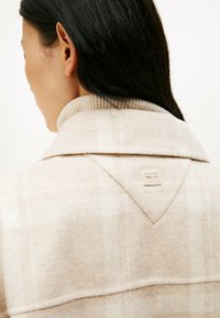 Beige plaid wool jacket featuring a ribbed collar and a triangular back yoke with a subtle embossed logo detail. Soft texture.
