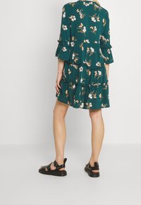 Abito floreale verde con maniche tre quarti, design a strati e dettagli arricciati. Abbinato a sandali neri con cinturini regolabili.