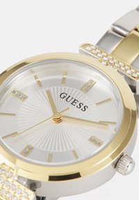 Montre en or et argent avec un boîtier rond, verre clair, cadran argenté avec motif éclat de soleil, accents dorés et marquages en cristal. Marque : Guess.
