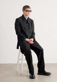 Trench coat negru cu dublă închidere, croi oversized, asortat cu pantaloni negri și pantofi masivi negri. Modelul poartă ochelari de soare negri.