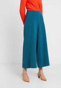 Turkosa vida byxor med hög midja och två sidofickor, tillverkade av mjukt tyg; kombinerat med en orange strukturerad långärmad tröja och beigefärgade klackar.