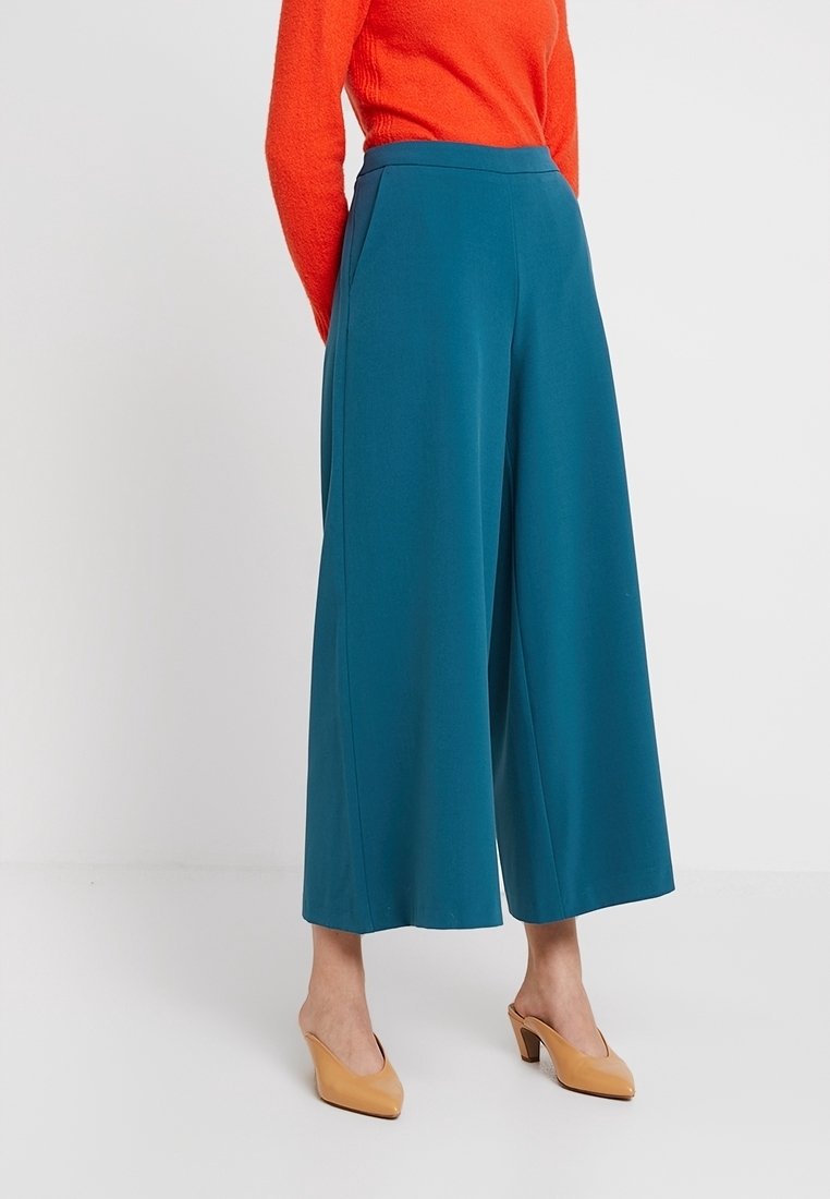 Turkosa vida byxor med hög midja och två sidofickor, tillverkade av mjukt tyg; kombinerat med en orange strukturerad långärmad tröja och beigefärgade klackar.