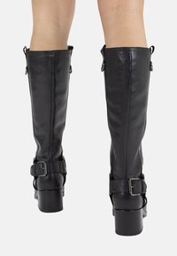 Bottes en cuir noir montantes, avec une finition lisse, présentant un large talon carré et des boucles réglables sur les côtés.