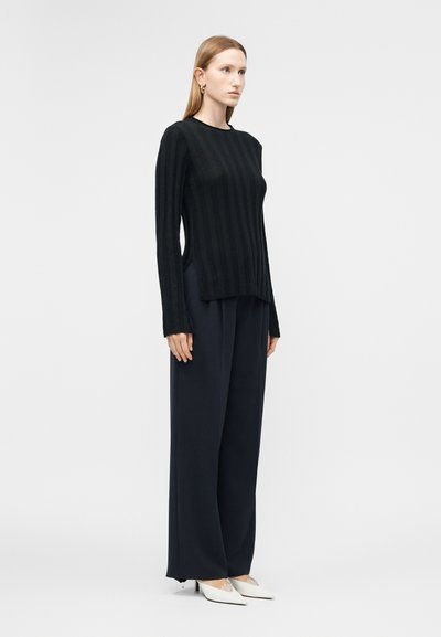 Femme en haut noir à manches longues, pantalon large bleu marine, et chaussures à talons blanches pointues, debout de profil sur un fond blanc uni.