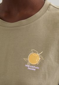 Olivengrünes Baumwoll-T-Shirt mit einem gelben Zitrusfrucht-Graphic und grünen Blättern sowie dem Text "Marco Polo Denim" in Lavendel.