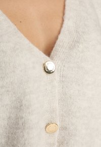 Cardigan gris clair en tissu doux et texturé, avec un col en V et trois boutons dorés sur le devant.