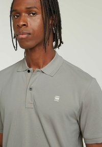 Szara koszulka polo wykonana z teksturowanej tkaniny, z trzema guzikami z przodu i małym białym logo na lewej piersi.