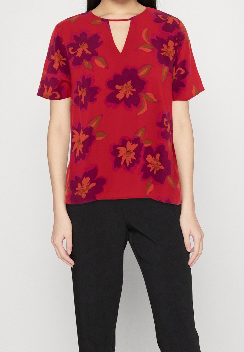 Rød bluse med korte ærmer, der har en nøglehulshalshalsudskæring og blomstermønster i lilla, pink og orange nuancer. Glat stoftekstur.