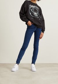 Černá mikina s grafickou potiskem, modré úzké džíny a bílé tenisky. Oblečení má volný střih a hladké textury.