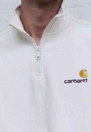 Gros plan d'une personne portant un sweat-shirt blanc à ouverture zippée quart de zip avec un logo Carhartt jaune brodé sur la poitrine.