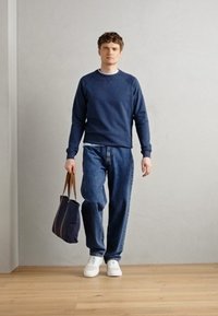 Sudadera navy con cuello redondo, jeans holgados y zapatillas blancas. Sosteniendo una bolsa tote navy con asas marrones. Diseño simple y casual.