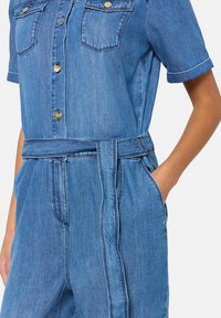 Salopette en denim bleu moyen avec des manches courtes, deux poches poitrine, une taille ceinturée et des coutures contrastées le long des coutures.