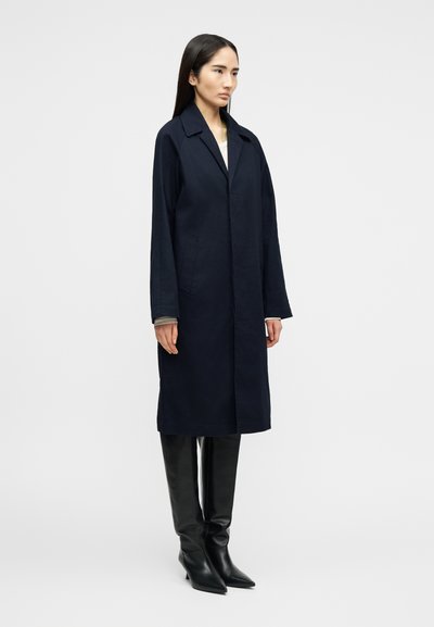 Γυναίκα που φοράει σκούρο μπλε παλτό μέχρι το γόνατο και μαύρες μπότες μέχρι το γόνατο, στέκεται μπροστά σε απλό ανοιχτό γκρι φόντο.