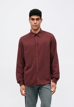 Mann, der vor einem schlichten Hintergrund steht und ein langärmliges, bordeauxrotes Hemd mit Knöpfen und verblasste graue Jeans trägt, und nach vorne schaut.