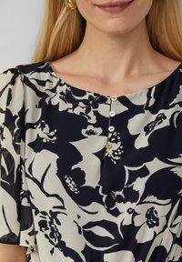 Fließende Bluse mit einem dunkelblauen Hintergrund und großen weißen Blumenmustern, Rundhalsausschnitt und fünf dekorativen Knöpfen vorne.