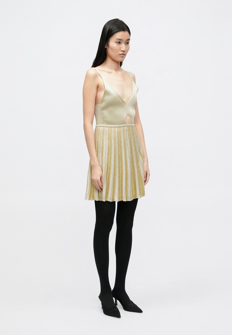 Goldfarbenes, metallisches Faltenkleid mit tiefem V-Ausschnitt und Cut-out-Detail, kombiniert mit schwarzen blickdichten Strumpfhosen und schwarzen spitzen Schuhen.