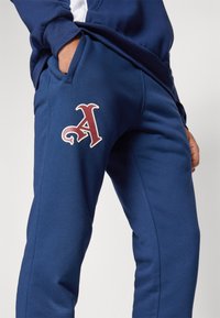 Temno modri trenirki z mehkimi vlakni, ki imata na stegnu poudarjen rdeč aplika z črko "A" ter stranske žepke.