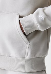 Ανοιχτό μπεζ φούτερ με ριμπ μανσέτες και ζώνη, το χέρι μερικώς μέσα στην μπροστινή τσέπη, μικρό κεντημένο λογότυπο στο μανίκι.