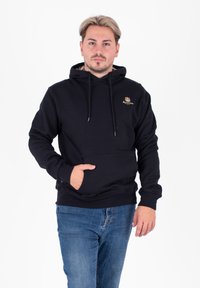 Felpa nera con cappuccio in misto cotone, con tasca frontale, coulisse e logo dorato sul petto. Indossata con jeans.