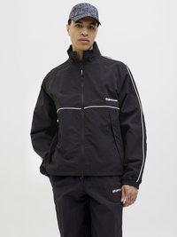 Black windbreaker jacket with white piping, high collar, and zip closure. Matched with black pants, featuring a logo and elastic cuffs.