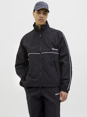 Veste coupe-vent noire avec des détails blancs, col haut et fermeture éclair. Assortie à un pantalon noir avec un logo et des poignets élastiques.
