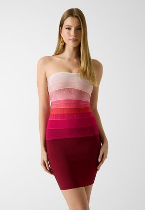 Kobieta o prostych blond włosach ubrana w sukienkę bez ramiączek, warstwową, ombre w odcieniach od jasnoróżowego do głębokiej czerwieni, stoi na jednolitym tle.