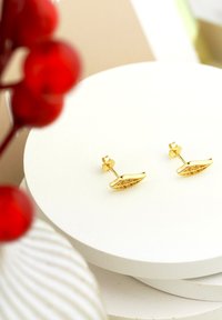 Guldörhängen i bladform med en texturerad design, visade på en rund vit pelare mot en mjuk bakgrund.