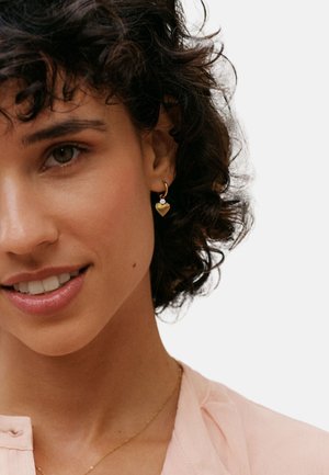 Guldfärgad hjärtformad hoops-örhänge med en liten accentsten, presenterad mot en neutral bakgrund. Modellen har lockigt hår och en ljusrosa topp.