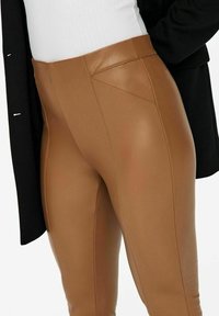 Hnědé umělé kožené legíny s hladkou texturou, se švy a vysokým pasem, zkombinované s černým vrchním oděvem.