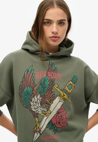 Hoodie vert olive avec un grand design brodé représentant un aigle, une épée et une rose, avec des couleurs vives et des motifs détaillés.