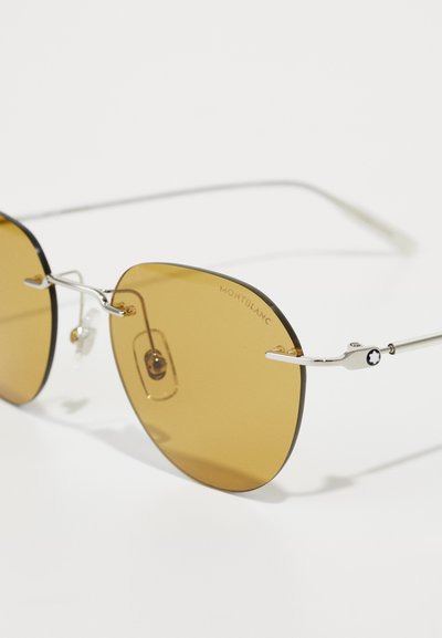 Lunettes de soleil rondes avec des verres marron, des montures en métal argenté et un petit logo noir et blanc sur la branche. Plaquettes de nez transparentes incluses.