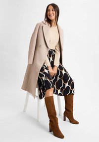 Frau, die auf einem weißen Stuhl sitzt, trägt einen beigen Mantel, einen gemusterten schwarz-weißen Rock, ein beiges Oberteil und braune kniehohe Stiefel.