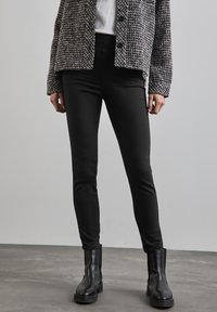 Zwarte skinny jeans met een gladde textuur, gecombineerd met een grijs-witte gestreepte jasje met knopen. Zwarte enkelboots maken de look af.