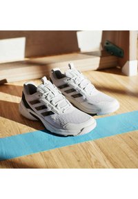 Białe buty sportowe z teksturowanym wierzchem, czarnymi akcentami i trzema czarnymi paskami. Gumowa podeszwa i sznurowana konstrukcja, na tle drewnianej podłogi.